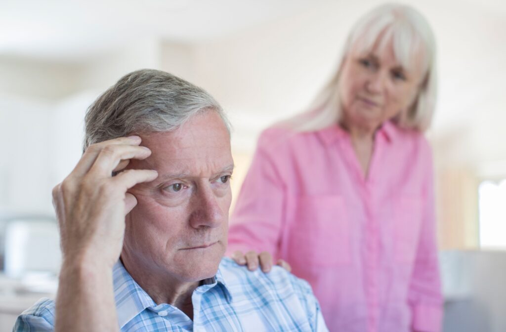 An older adult touches their head with their right hand to remember something they forgot while their spouse watches in concern